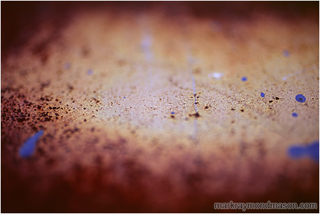 Abstract photograph of blue paint splatter seeming to hover above a blurry, rusted steel plate