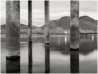 Concrete Piles, Rushing Water: Kamloops, BC, Canada (2012) - Fine art black and white photograph of large piles rising out of the frame from the smooth water of a full river