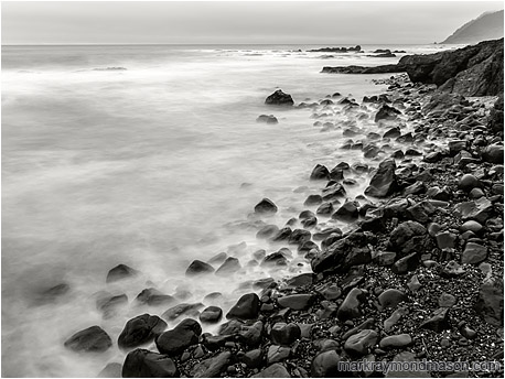 Long exposure black and white photo of water crashing and receding on a beach littered with shiny rocks