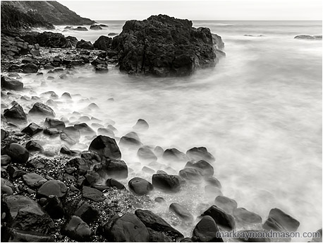 Fine art black and white photograph of smokey waves swirling around a rocky knob and a bounder-strewn beach