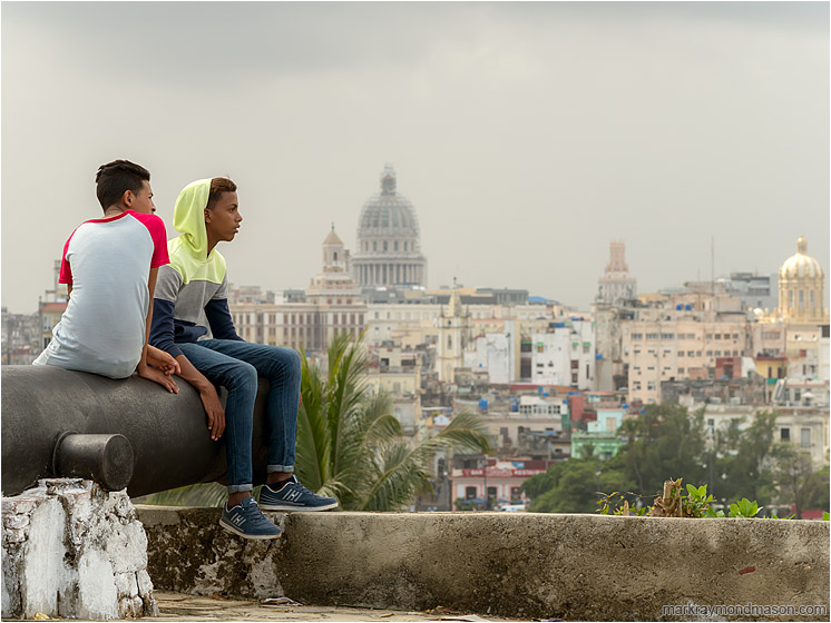 Two Boys, Cannon, Skyline: Havana, Cuba (2017-02-16) - Fine art photograph showing two teenage boys sitting on an ancient cannon, the Havana skyline and grey skies in the background
