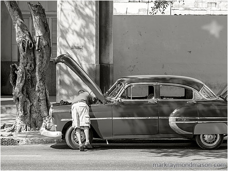 Fine art black and white photo showing a man bending into the open hood of a finely restored classic car