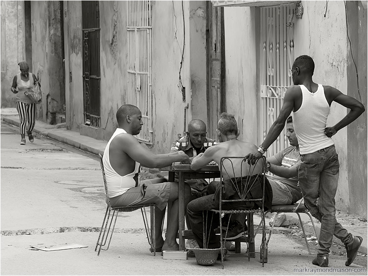 Dominoes Game, Walking Woman: Havana, Cuba (2017-02-19) - Fine art black and white photograph showing a group of men playing dominoes at a table in the street, and a woman with a cigarette and shopping bag in the background