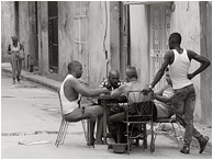 Dominoes Game, Walking Woman: Havana, Cuba (2017) - Fine art black and white photograph showing a group of men playing dominoes at a table in the street, and a woman with a cigarette and shopping bag in the background