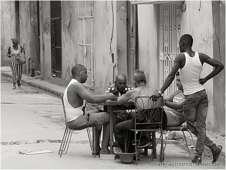 Fine art black and white photograph showing a group of men playing dominoes at a table in the street, and a woman with a cigarette and shopping bag in the background