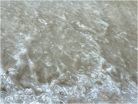 Abstract photo of cascading bubbles frozen in place in the thick, cracked ice of an urban stream