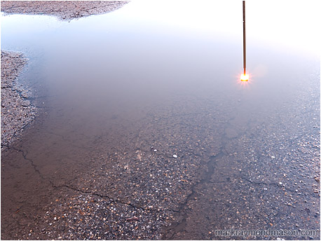 Fine art photograph showing a lamp post reflected in a pool of water on cracked pavement