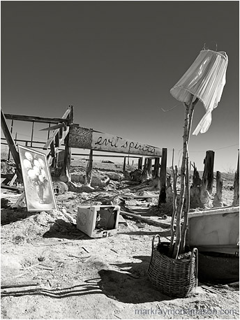 Fine art B&W photograph showing the remains of a structure and scattered objects on a salt flat