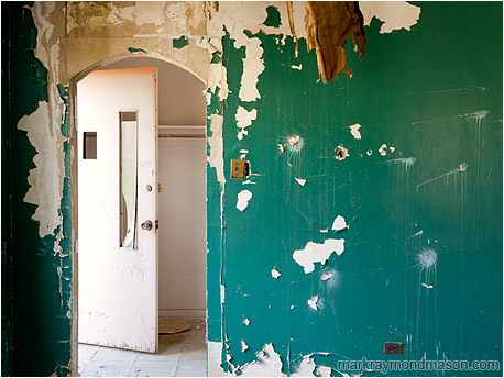 Fine art photograph of an archway and a slack door, set behind a peeling, pitted, bizarrely painted wall