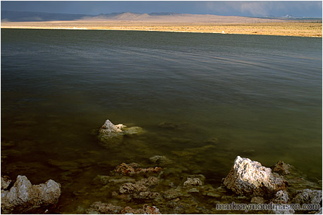 Fine art photograph of small volcanic rocks in a large lake, with stormclouds and sunshine in the distance
