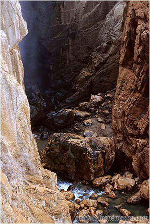Fine art nature photograph of water and mist in a deep slot canyon, with steep cliffs of orange and black rock