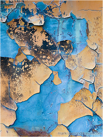 Abstract macro photograph showing large chunks of peeling orange paint falling away to reveal cool, blue-tinted metal beneath