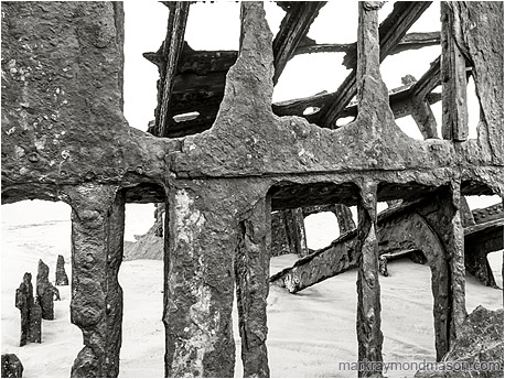 Fine art black and white photograph of the metal skeleton of an old wrecked ship, surrounded by pale, ocean-worn sand.
