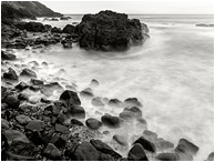 Worn Rocks, Crashing Waves: Near Stawberry Hill Park, OR, USA (2015) - Fine art black and white photograph of smokey waves swirling around a rocky knob and a bounder-strewn beach