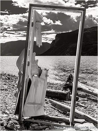 Fine art black and white photograph of a disembodied standing door with no walls and a beautiful background of lake, sky and mountains