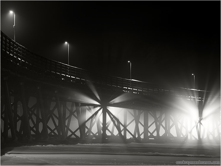 Lamplight, Crossed Beams, Fog: Salmon Arm, BC, Canada (2016-12-30) - Fine art black and white photo showing light streaming through the crossed beams of an ice-locked wharf in the night