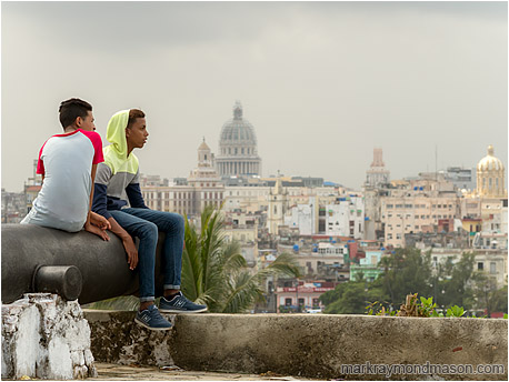 Fine art photograph showing two teenage boys sitting on an ancient cannon, the Havana skyline and grey skies in the background