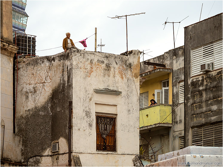 Man, Woman, Concrete Houses: Havana, Cuba (2017-02-16) - Fine art photograph showing a man perched unsettlingly on top of a concrete building and a woman looking into the street from a mid-level apartment
