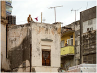 Man, Woman, Concrete Houses: Havana, Cuba (2017) - Fine art photograph showing a man perched unsettlingly on top of a concrete building and a woman looking into the street from a mid-level apartment