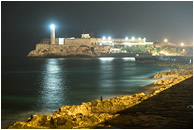 Tower, Ocean, Distant Figure: Havana, Cuba (2017) - Fine art photograph of a figure on the rocks by the sea, with floodlights on a fortress and a lighthouse in the background