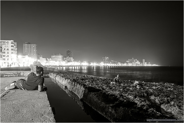 Resting Man, Fishing Party, Skyline: Havana, Cuba (2017-02-17) - Fine art black and white photograph of a man resting on the seawall in Havana, taking in the city lights and a party fishing by lamplight