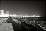 Resting Man, Fishing Party, Skyline: Havana, Cuba (2017) - Fine art black and white photograph of a man resting on the seawall in Havana, taking in the city lights and a party fishing by lamplight