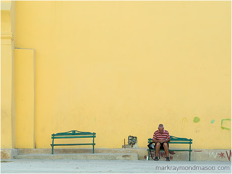 Fine art travel photo of a man seated alone on a bench in front of a towering orange wall