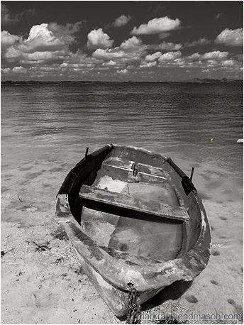 Fine art black and white photograph showing a flooded boat, cocked sideways, sitting abandoned on the silty shores of a shallow lake