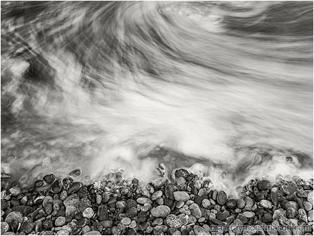 Fine art black and white long-exposure photograph showing moving water swirling like fog around seaside stones