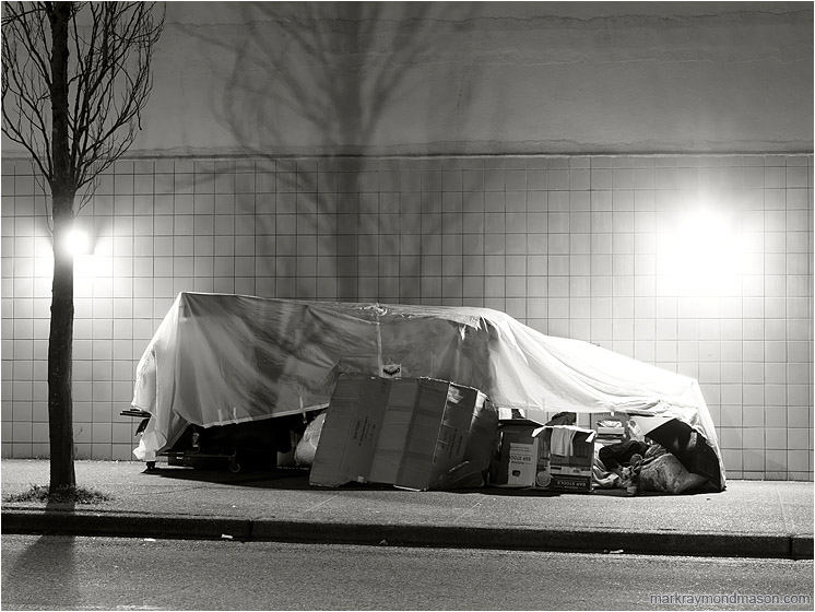 Street Shelter, Sleeping Figure: Vancouver, BC, Canada (2018-03-20) - Black and white photograph of a homeless person sleeping in a crude tarp and cardboard shelter against a tile wall
