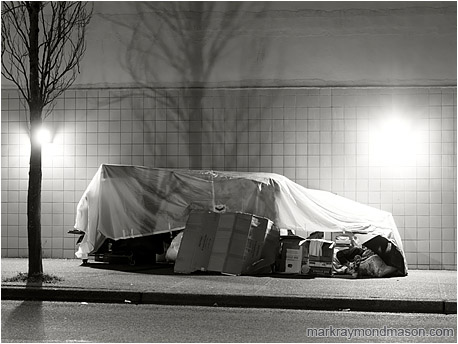 Black and white photograph of a homeless person sleeping in a crude tarp and cardboard shelter against a tile wall